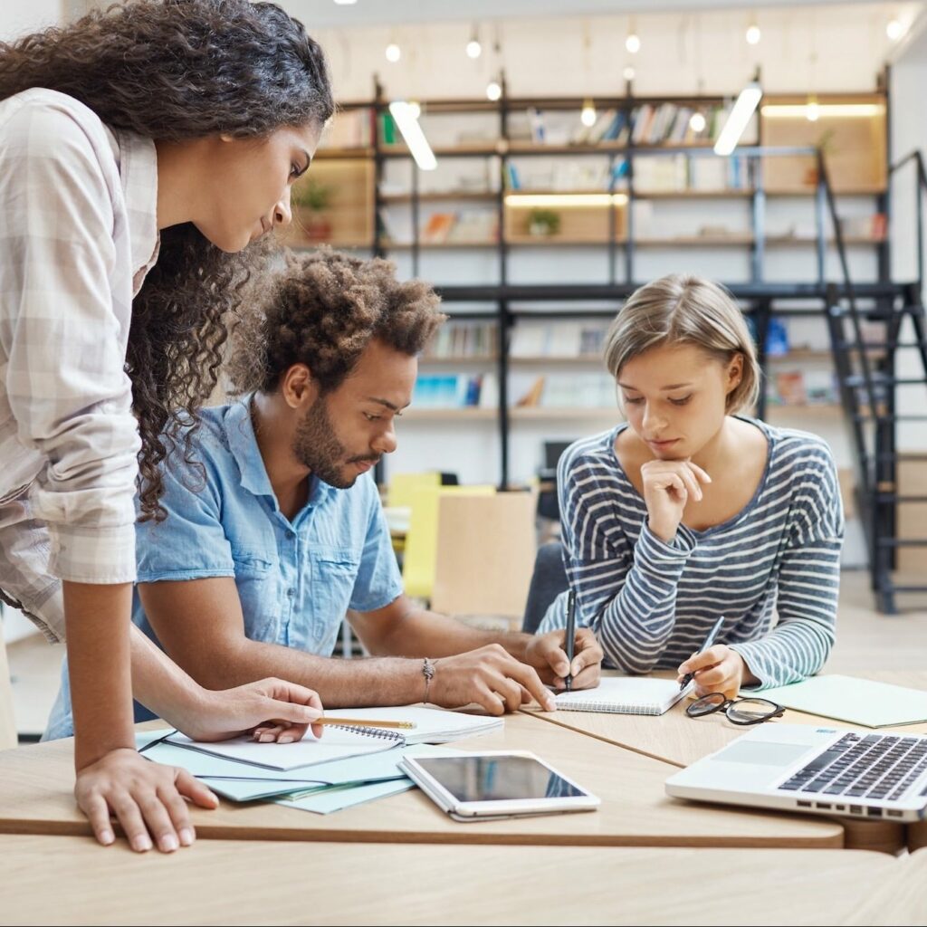 Deux personnes assises suivant une formation en management de projet en entreprise accompagné par le formateur se penchant au dessus d'eux. 